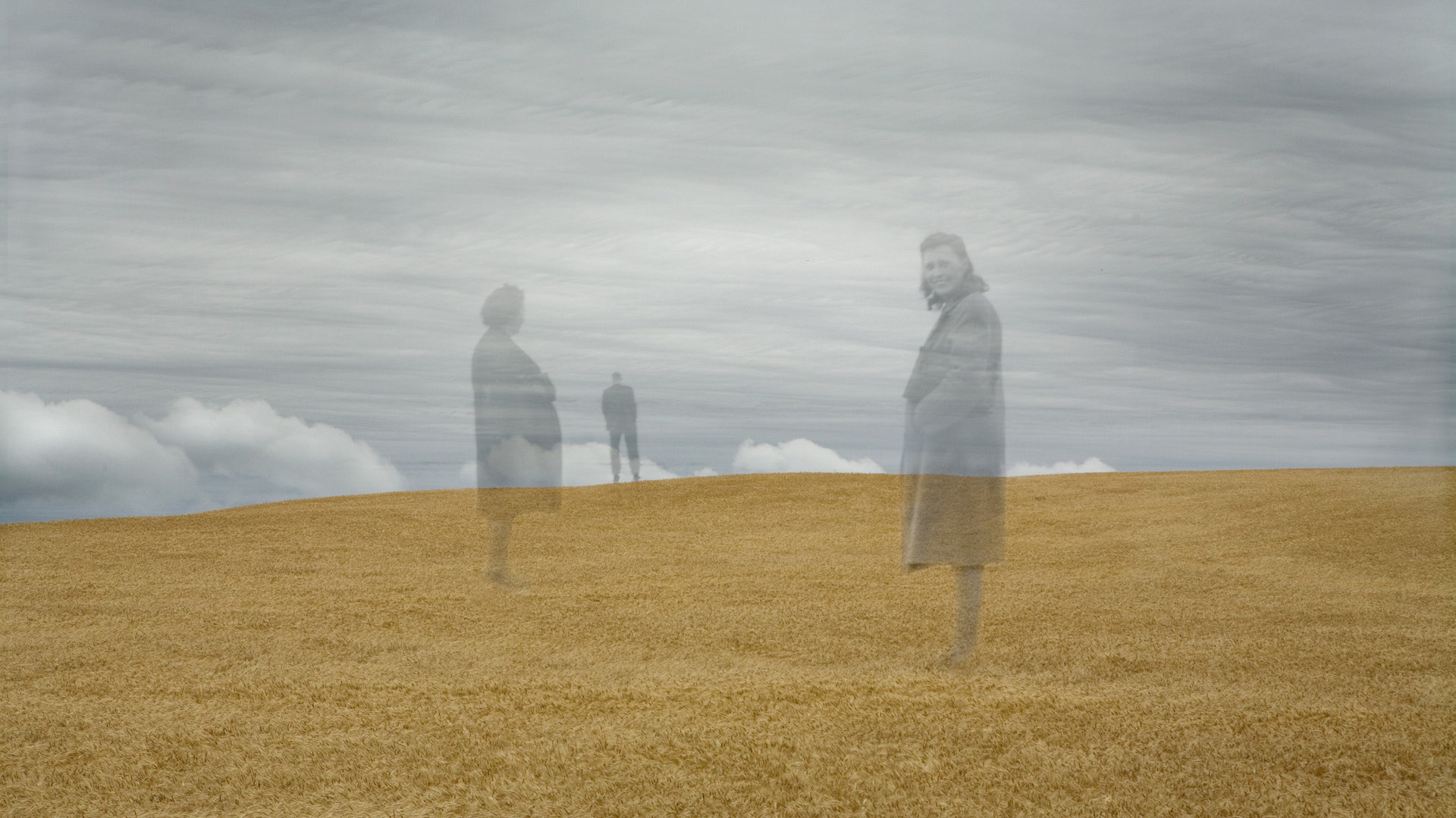 Three people from a black and white photo, overlayed on a recent color photo of a wheat field with gray skies in the background