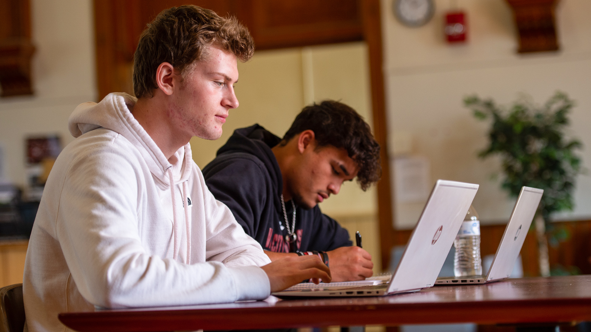 2 male students sit at a table while doing classwork on 2 computers