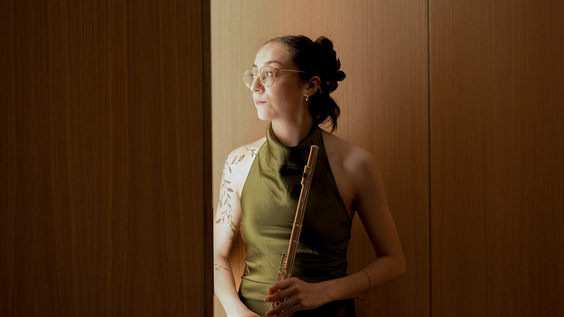Ela Day Child, wearing a green dress, holds a flute while leaning against a shadowed wood wall