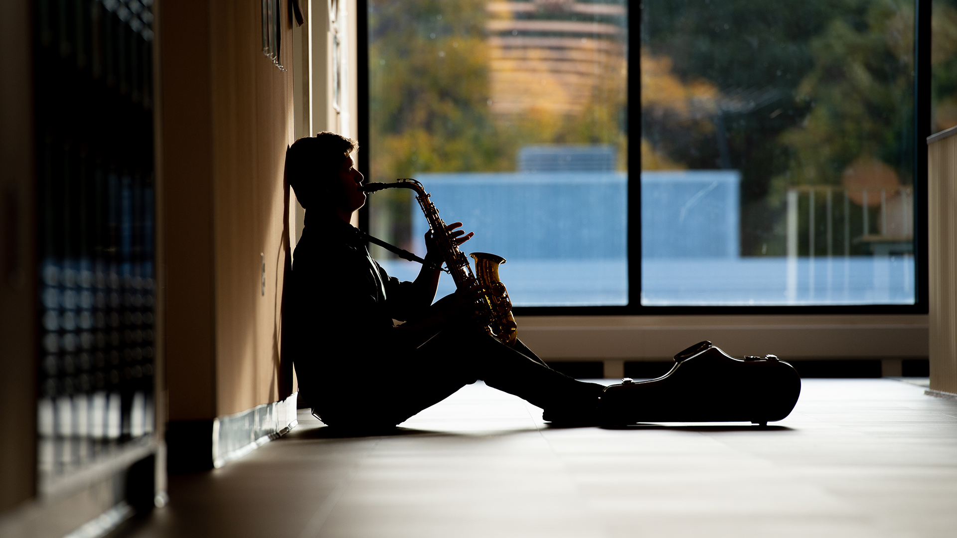 Male student playing saxophone, sitting on floor in hallway