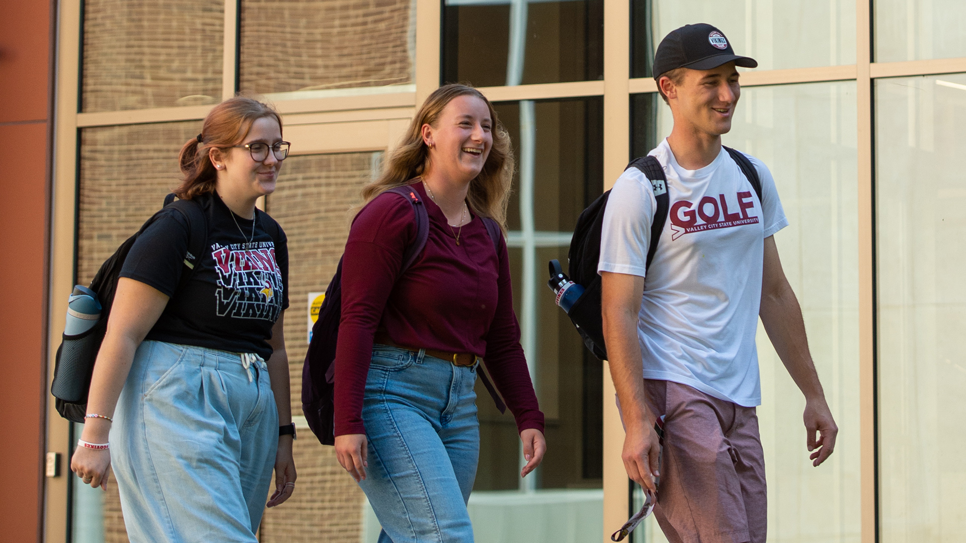 One male and two female students walk on VCSU's campus in front of glass windows