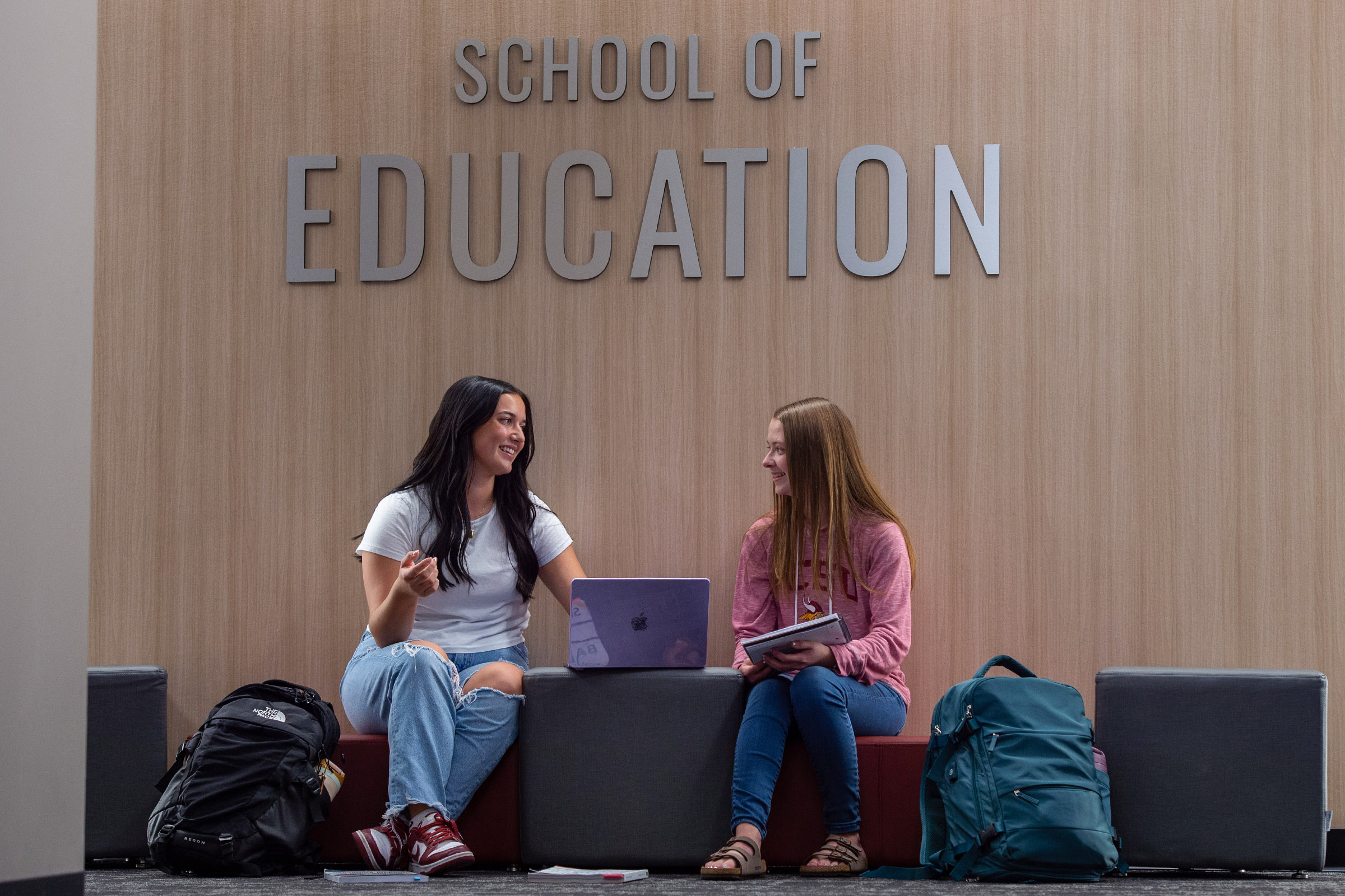 Two students sit work on a laptop in front of a sign that says School of Education