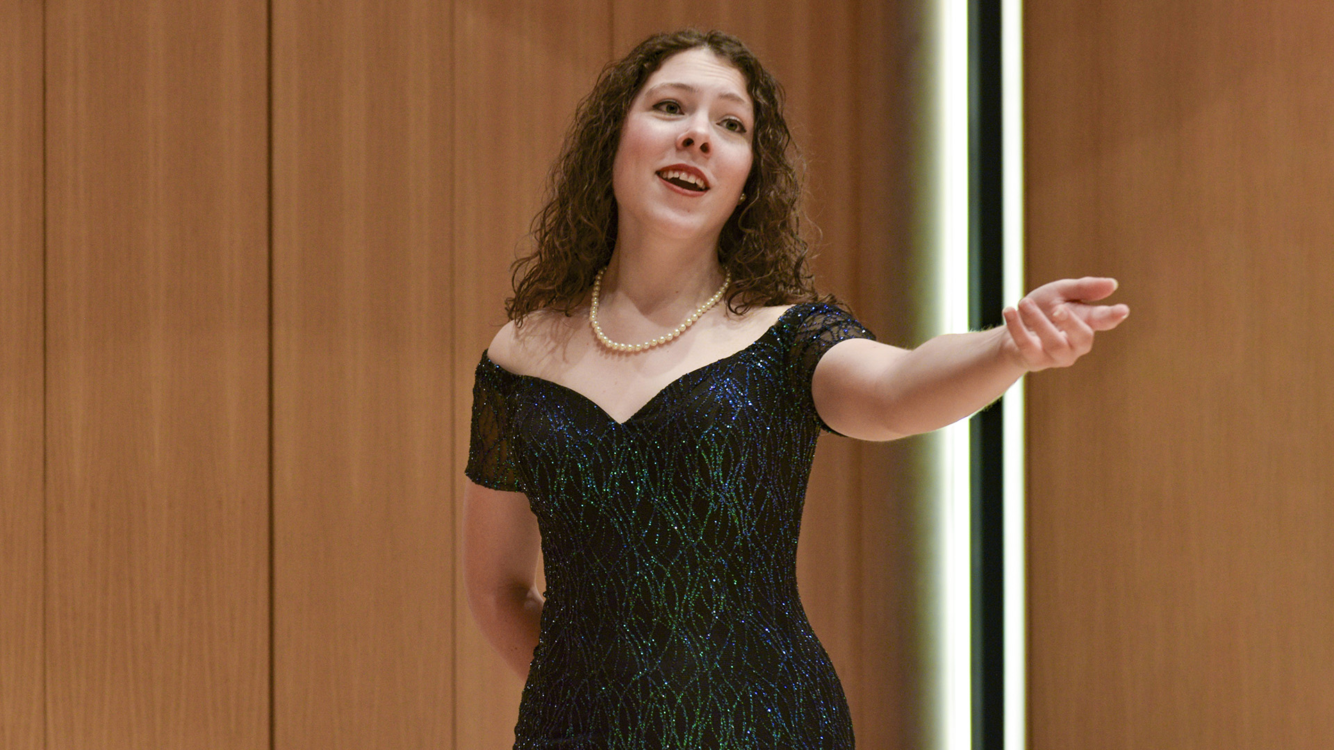 Elizabeth Gazeley, wearing a green and black dress, reaches her arm out as she sings from the stage at the Center for the Arts.