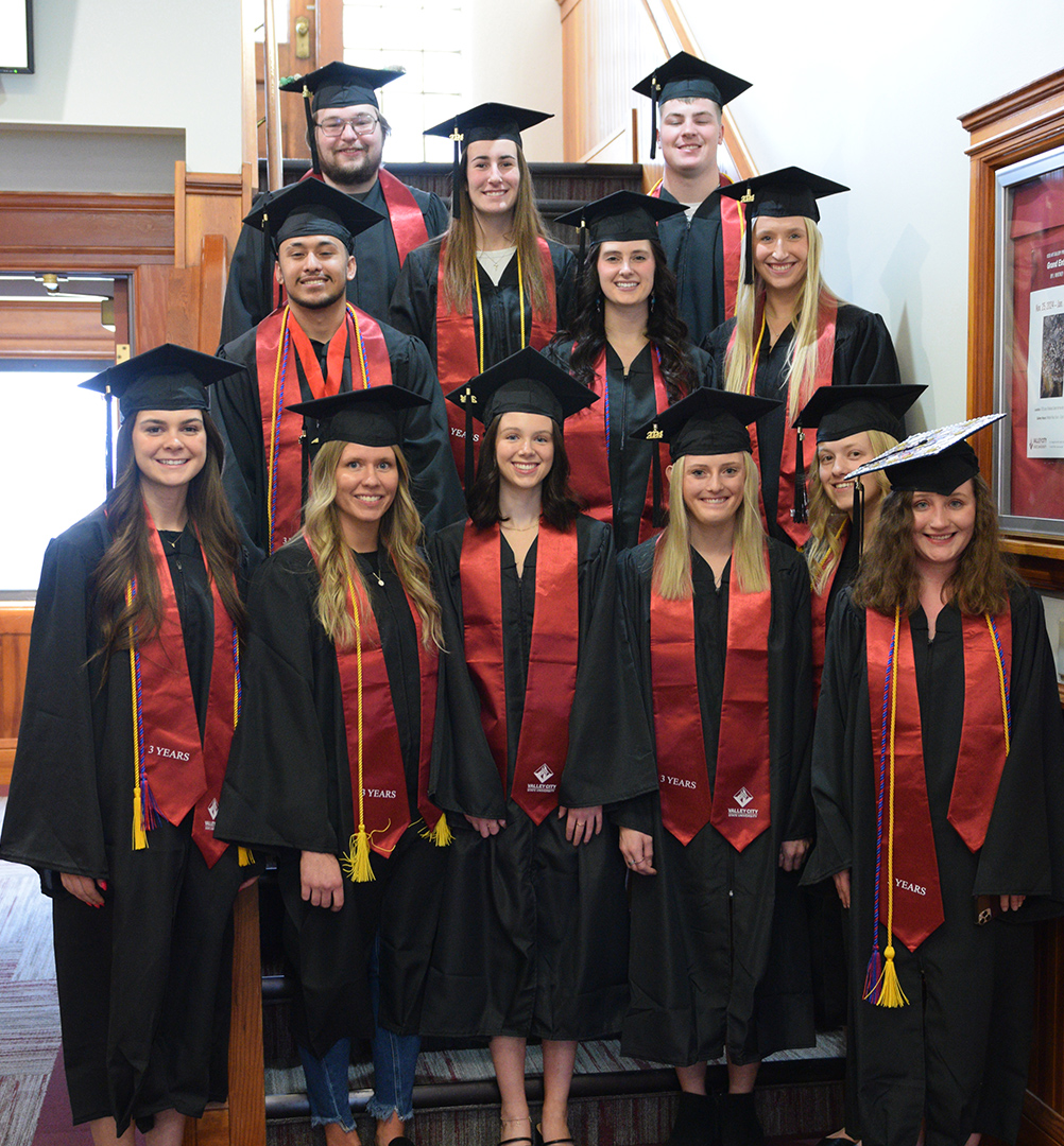 VCSU's 12 three-year graduates wear bright red stoles along with caps and gowns while standing together on stairs.