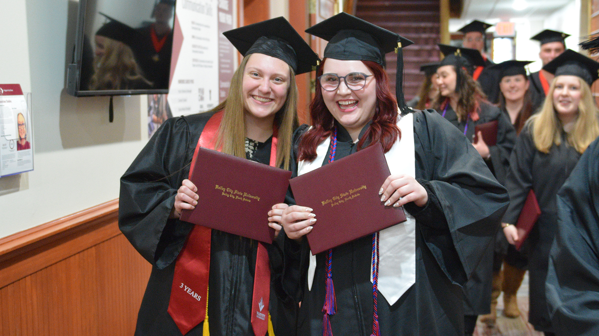 2 VCSU graduates hold up their diplomas while smiling
