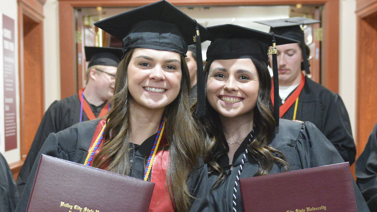 Two VCSU students in graduation caps and gowns smile while holding up their VCSU diplomas