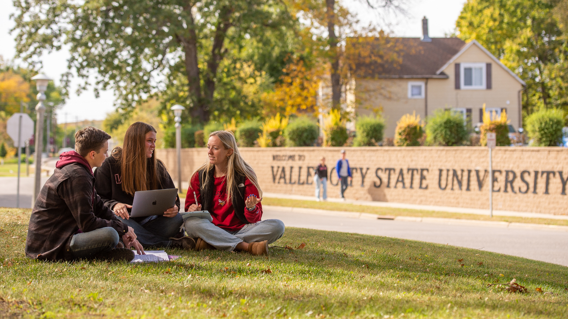 3 VCSU students sit on the lawn in front of a sign saying Valley City State University