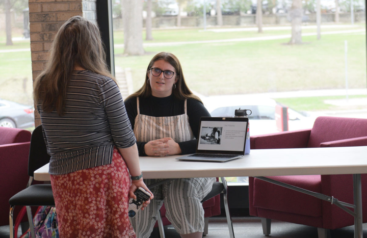 student siting at table, presenting to person whose back is facing camera. Laptop on table and greenery outside of the window behind student