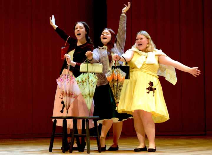 Three students in poodle skirts, singing with arms raised and purses and umbrellas in hands