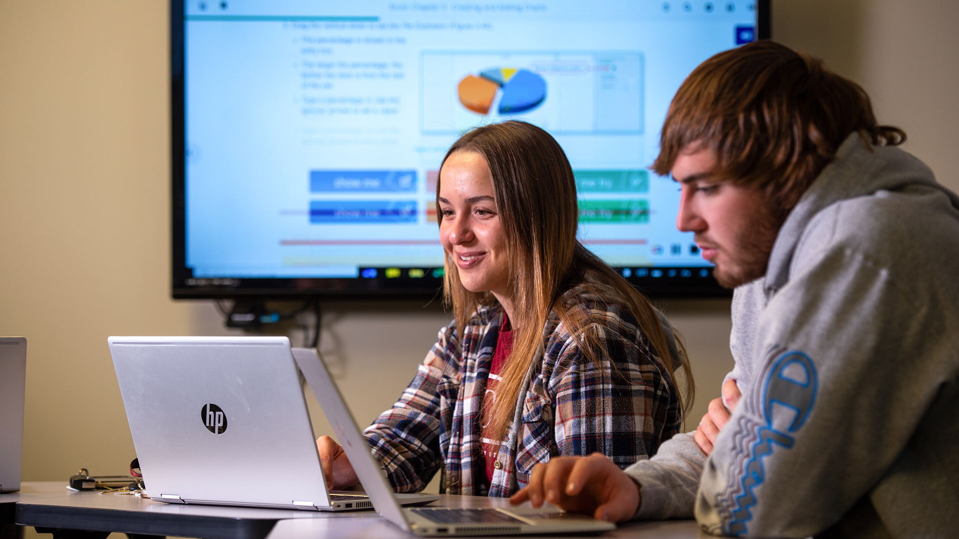 2 VCSU business students work on computers with a presentation on a screen in the background