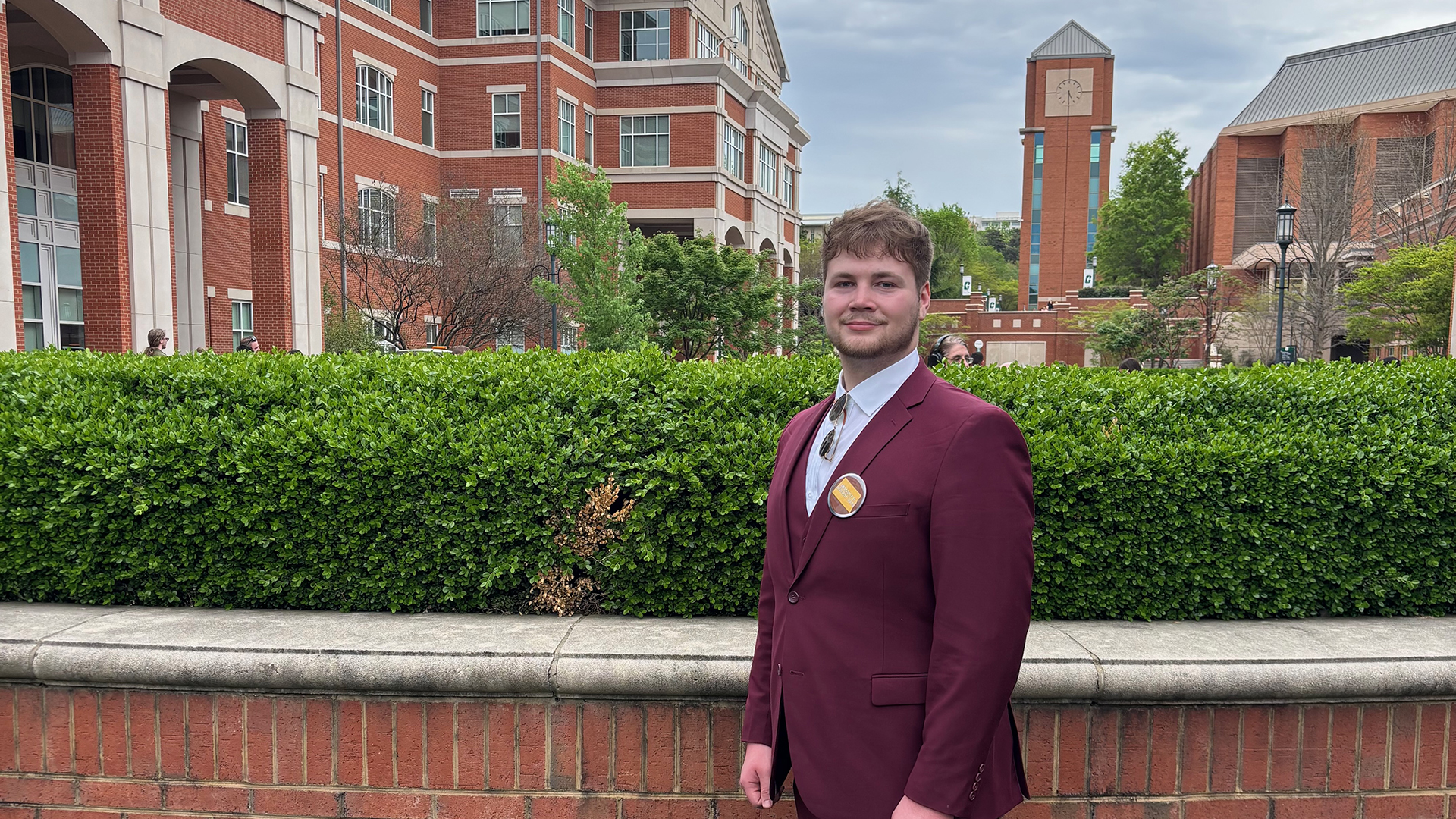 Grady Hornung, wearing a deep red suit, stands in front of brick buildings on the UNC-Charlotte campus.