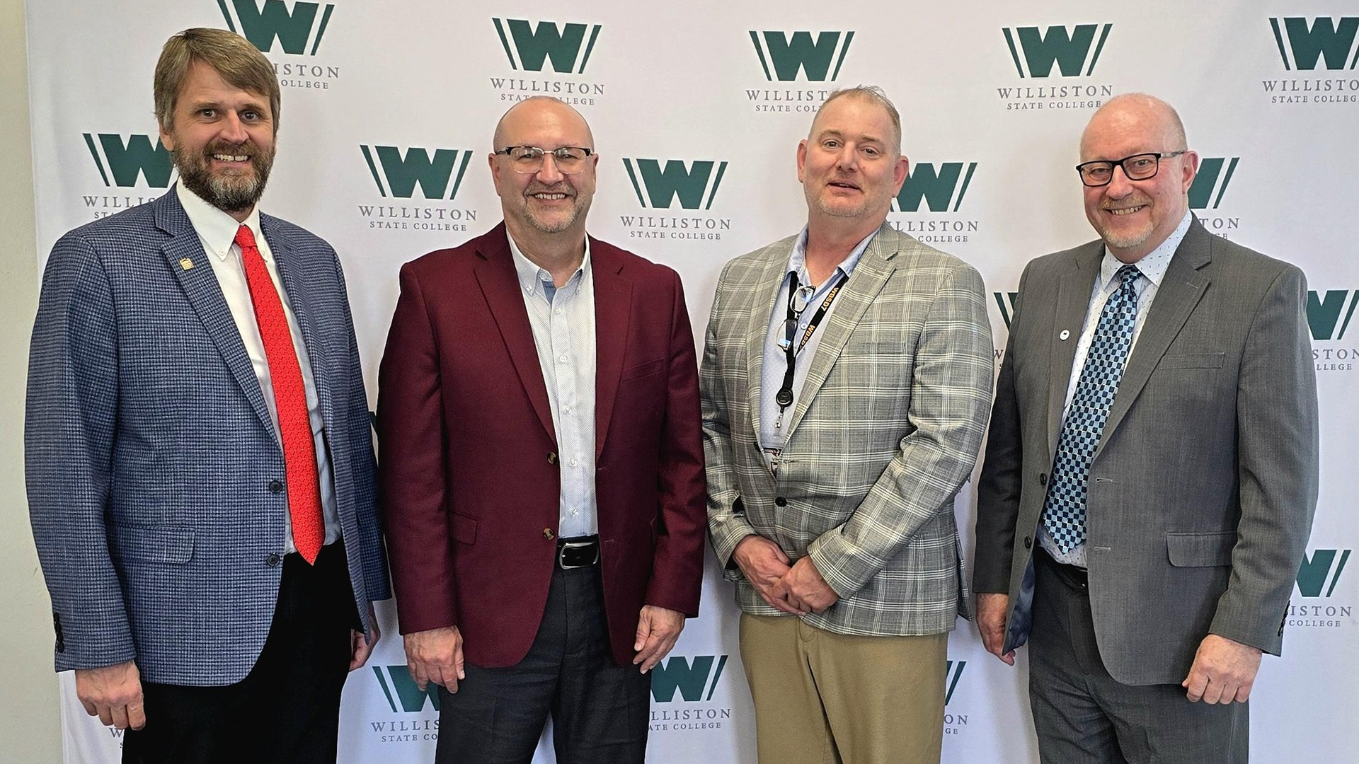 Four men in suits stand in front of a white Williston State College backdrop