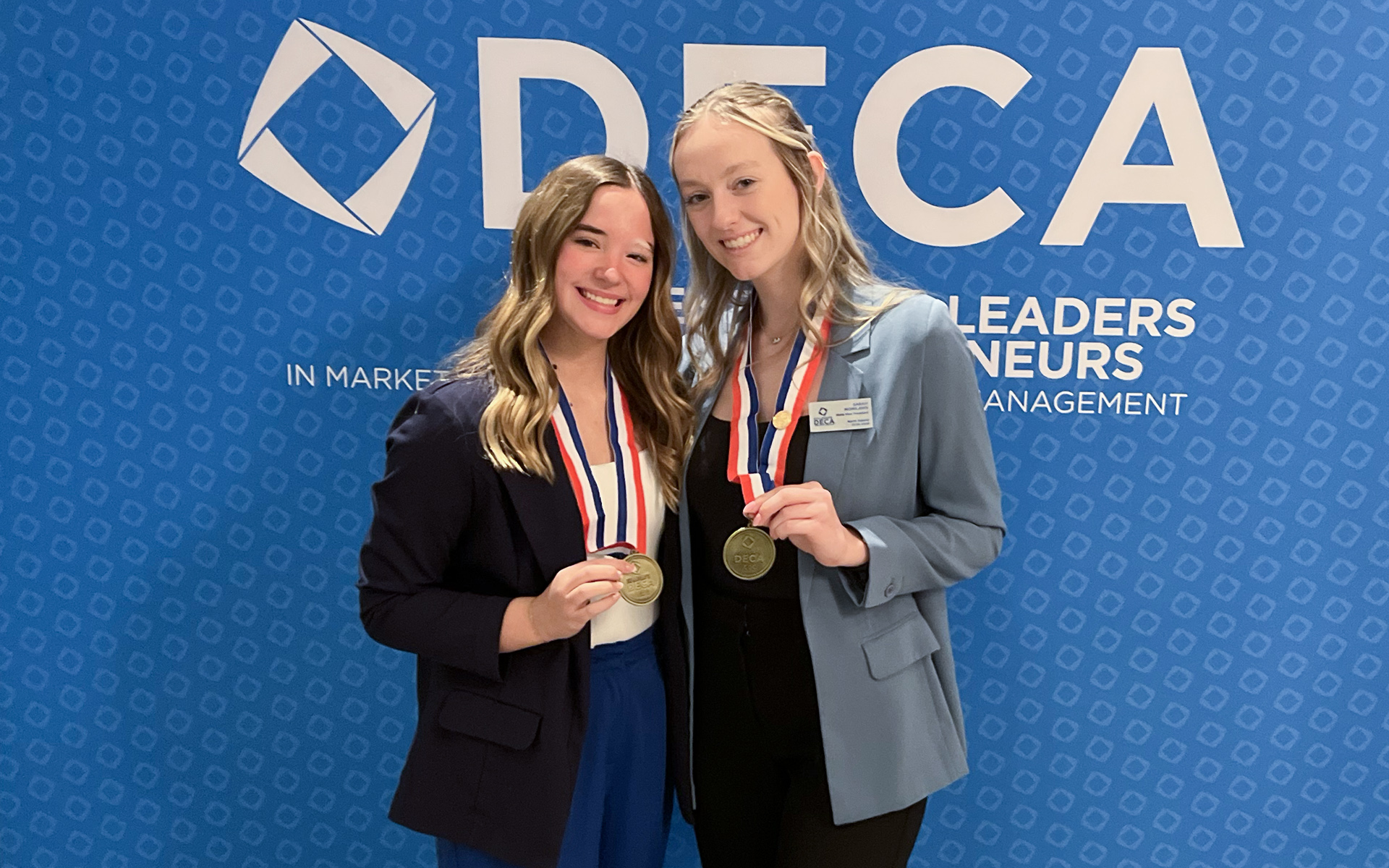 Katie Jones and Sarah Monilaws are pictured in professional dress, wearing medals in front of a blue DECA backdrop