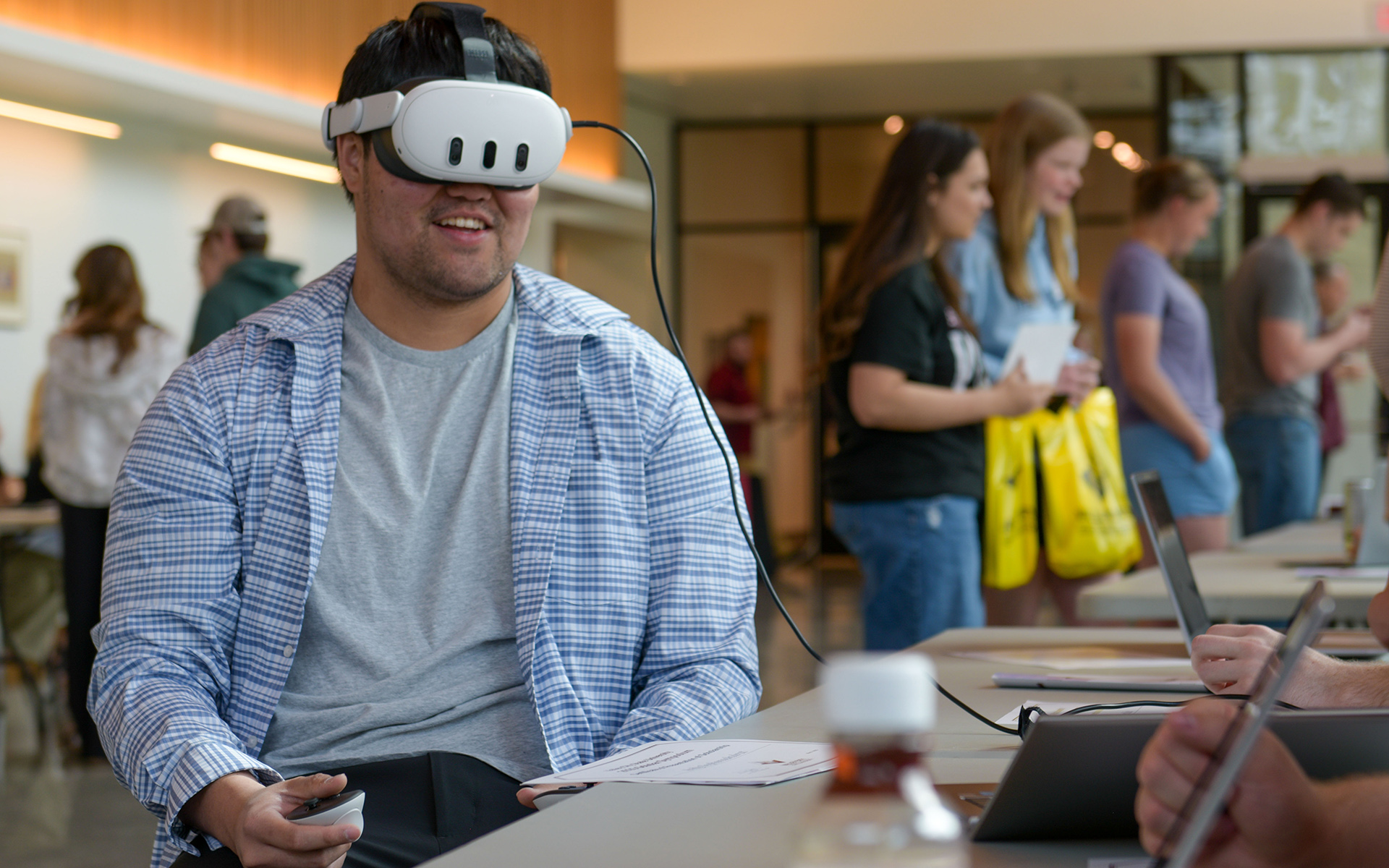 A male student wears virtual reality goggles during the 2025 Scholar Symposium as he experiences a video game developed by a VCSU student. Other students interact at tables in a large lobby.