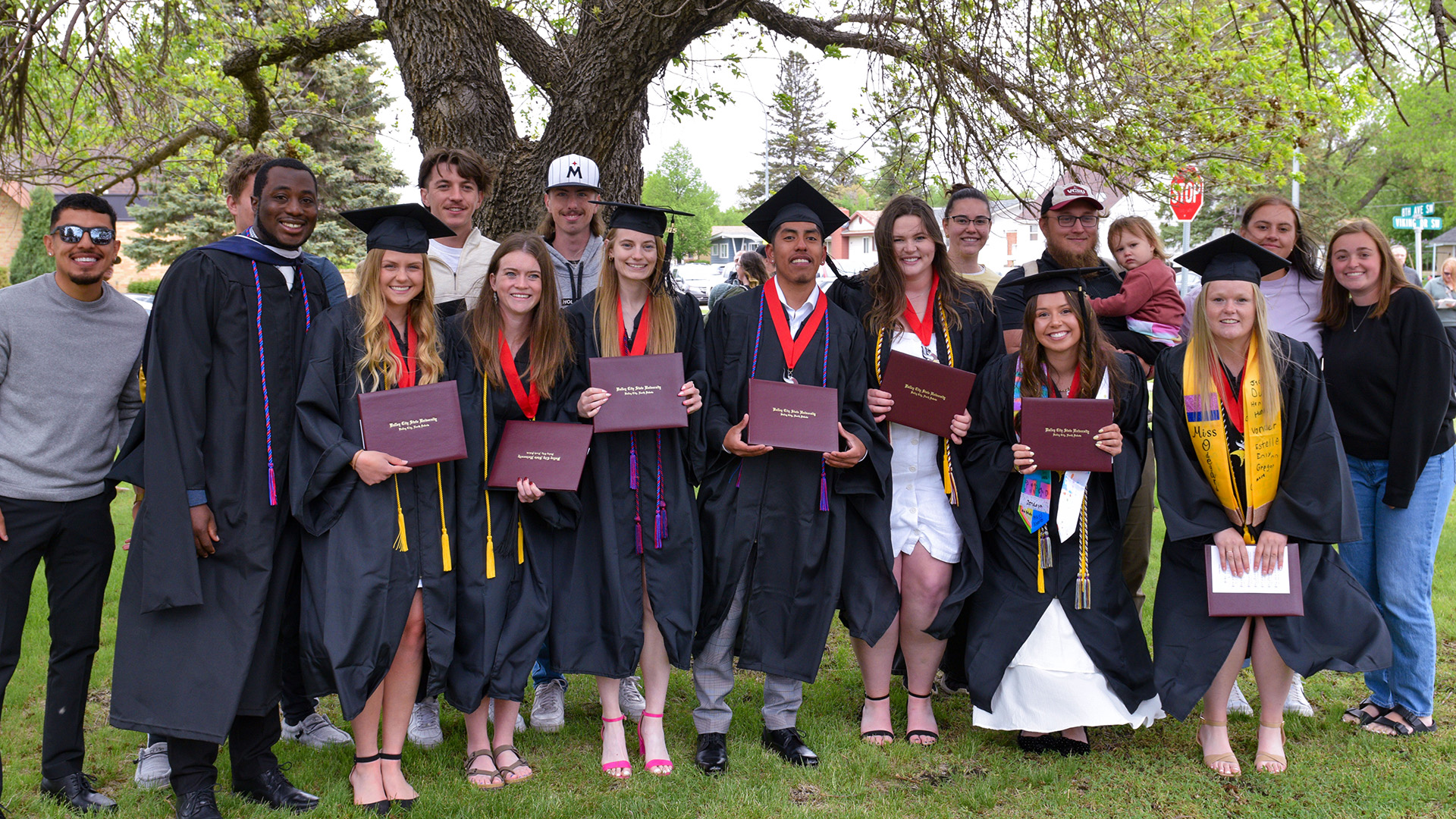 A group of VCSU graduates in black cap and gown smile with their friends while holding diploma covers.