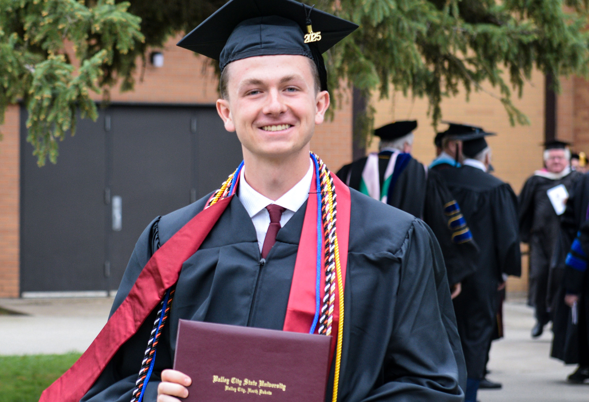 Grant Larson, wearing a black hat and cap and a red stole, smiles for the camera while holding his diploma cover.