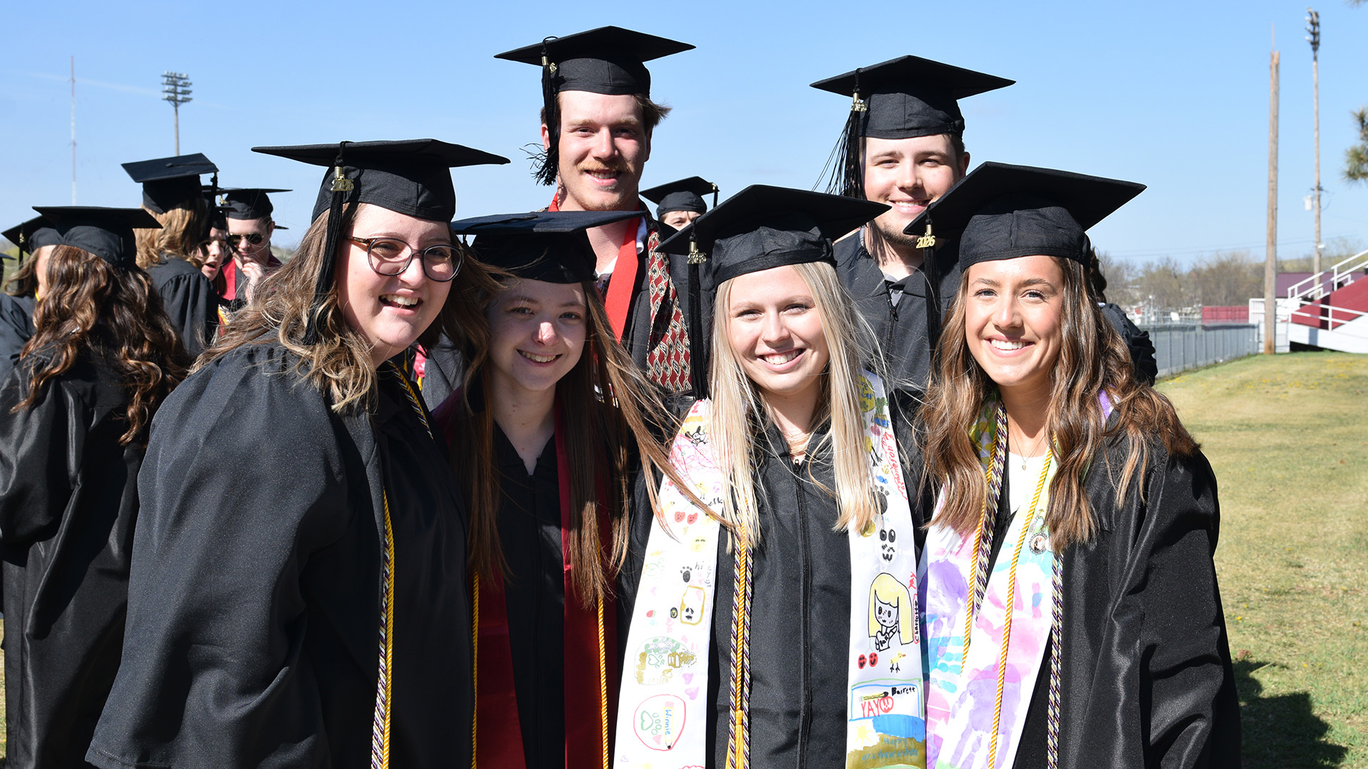 Several VCSU graduate in black robes and caps pose for an outdoor smiling photo prior to commencement.