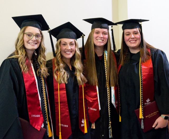 group of four female graduates in cap and gown smiling at camera wearing three year stoles