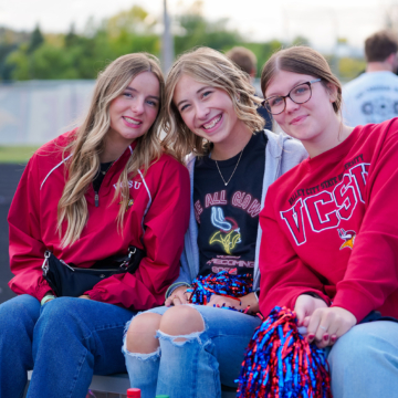 three students smiling, sitting together with pom poms outdoors, wearing VCSU attire