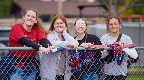 4 students leaning against chainlink fence, holding pom poms, smiling at camera