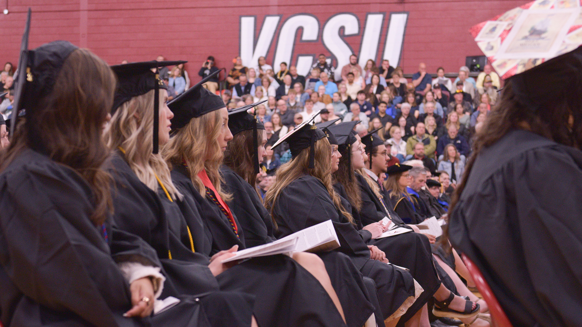 VCSU students wear black graduation gowns and caps, sit in red chairs and listen to a speaker at commencement