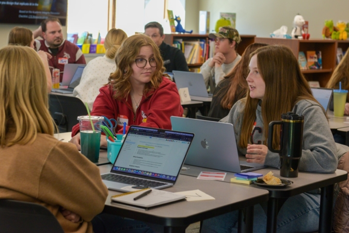 two students talking at a table with other people
