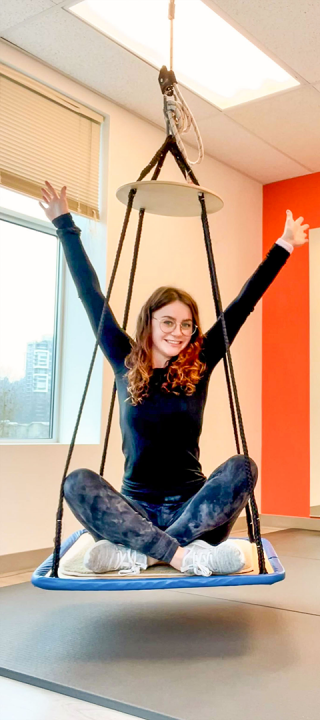 Darrien Cantelo with arms raised above her head, sitting cross-legged in a swing in her physical therapy studio.