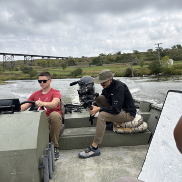 Student driving a boat on the Sheyenne River while a man with a camera films him