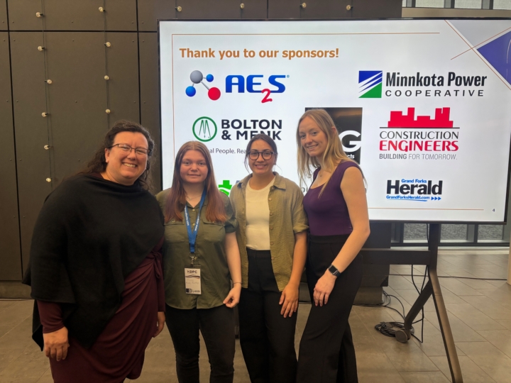 Tammy Jo, Trinity, Hanna and Sarah standing in front of sign with conference sponsors listed