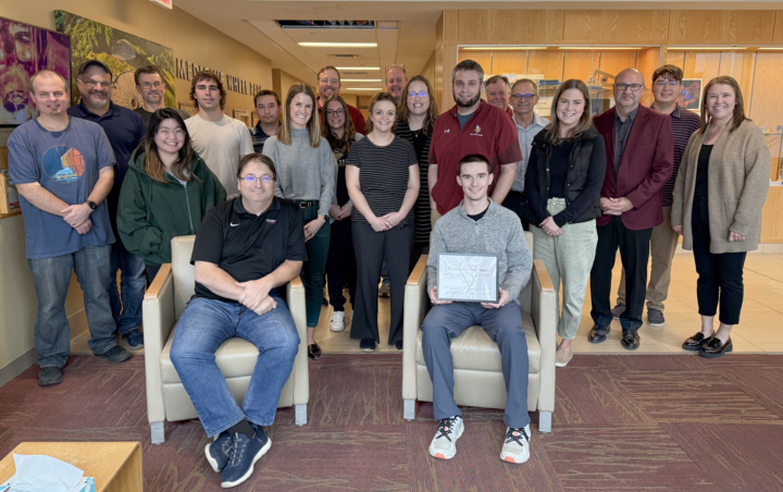 Group of people with two sitting in chairs in the foreground and multiple others standing behind smiling at the camera