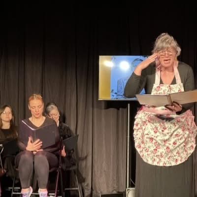 Jenni Lou performing on stage with apron on and others sitting in background
