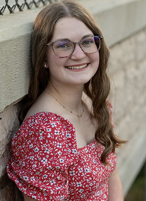 Kristyn Bauer wears a red and white dress for a senior photo as she leans against a brick wall
