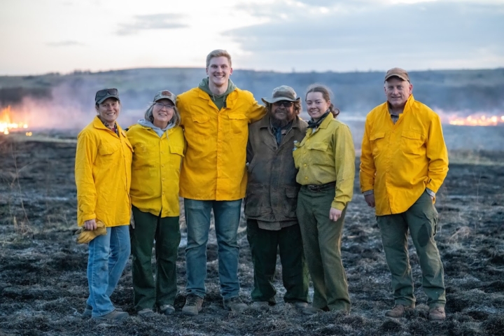 Six people wearing yellow work gear with fire in the back