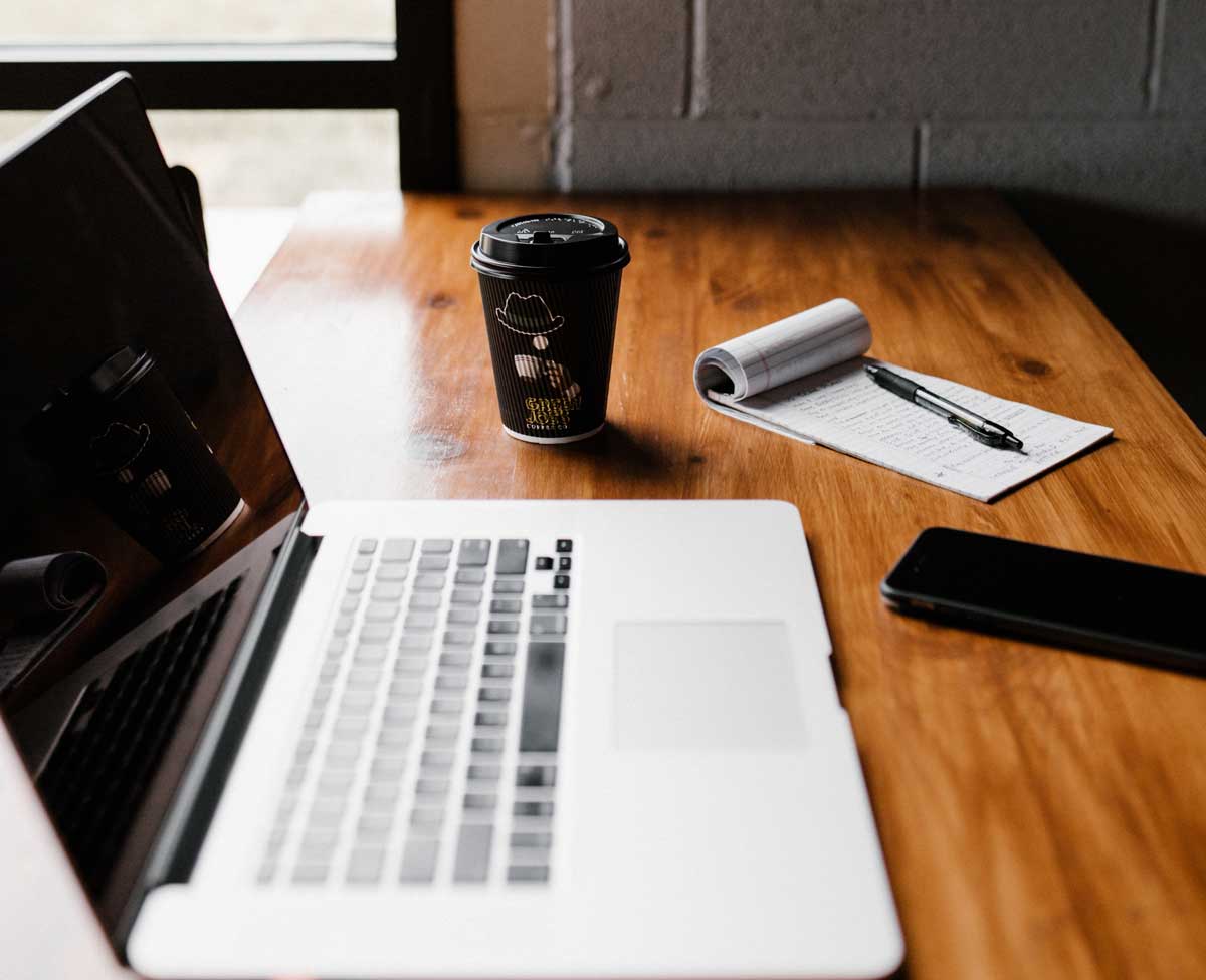 A picture of a laptop computer, coffee and a pen and paper on a desk.