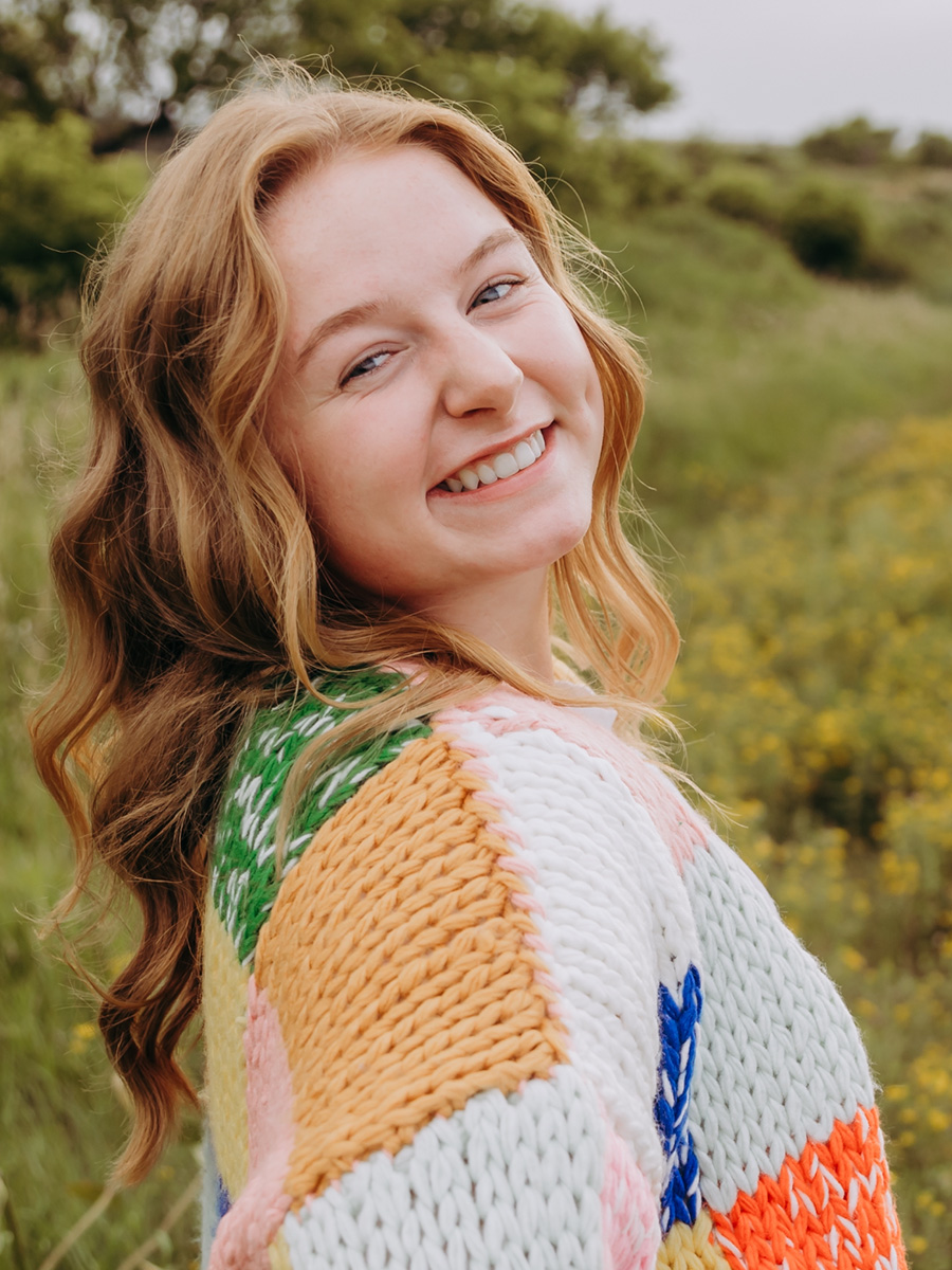 Rhyan Jenson wears a colorful sweater and smiles for the camera in front of a background of green grass and trees