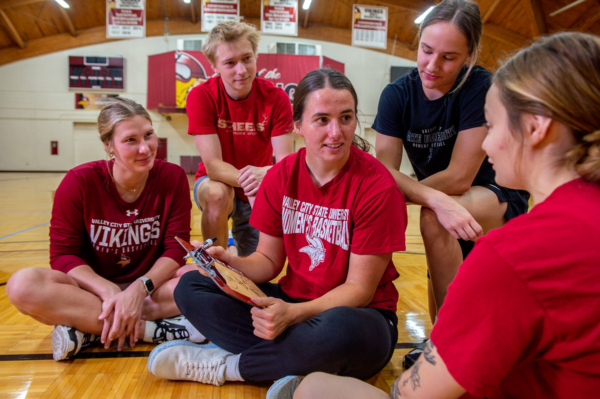 Four students sit and listen to a coach on the floor of the Bubble fieldhouse.