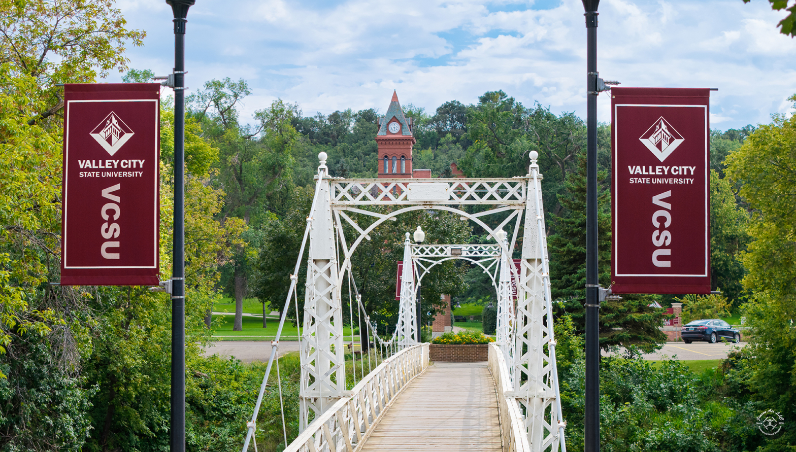 The VCSU walk bridge is pictured, flanked by two VCSU banners, and the McFarland Hall clocktower in the background. Photo by Jensen Air LLC.