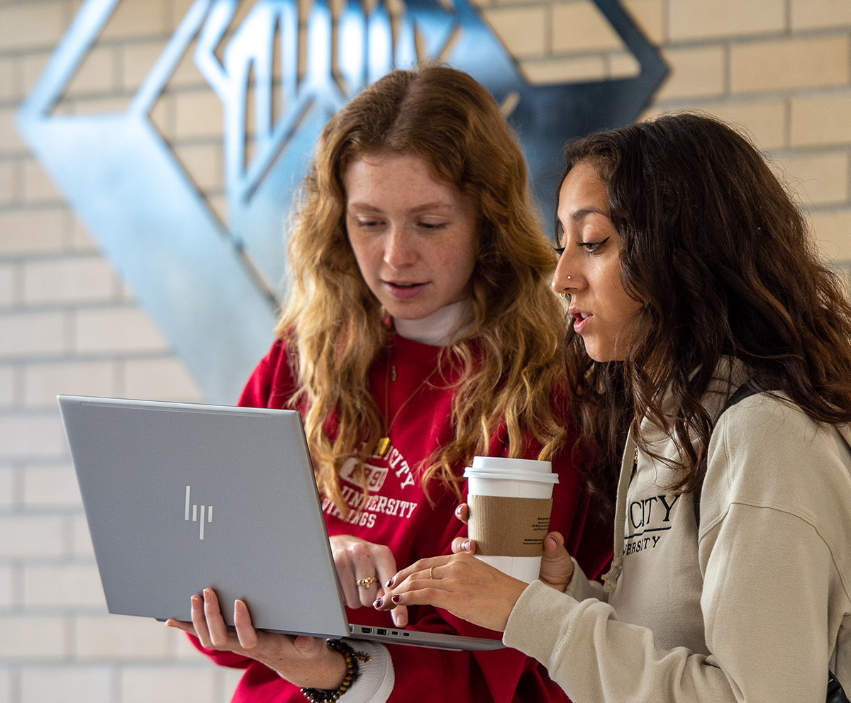 two female students standing side by side, looking at laptop and having conversation