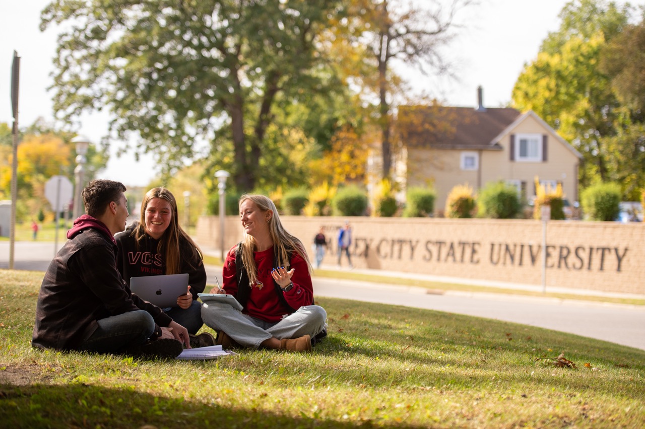 Three VCSU students sitting on grass in the foreground, with a wall in the background inscribed with Valley City State University in large letters.