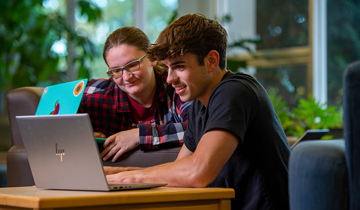Two students sit at a desk and look at an open laptop