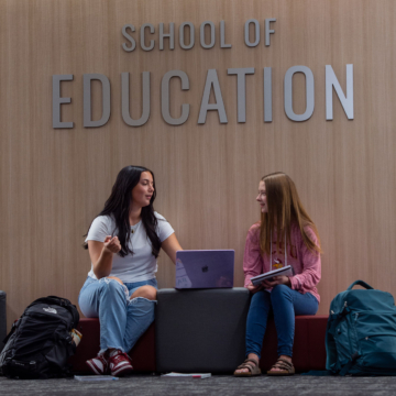 Two students sit work on a laptop in front of a sign that says School of Education