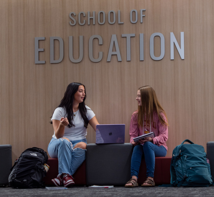 Two students sit work on a laptop in front of a sign that says School of Education